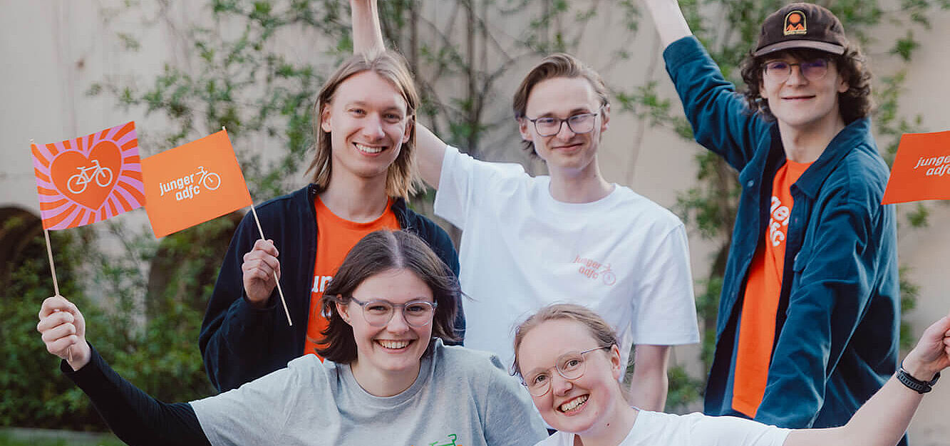 Fünf junge Menschen posieren draußen auf und neben einem Lastenrad. Sie lachen und halten kleine Fahnen des Jungen ADFC in die Höhe.