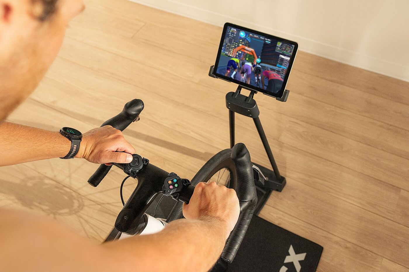 Tacx Bildschirm Hände liegen an einem Rennradlenker. Rechts steht ein Tablet auf einem Ständer, auf dem eine Gruppe Radfahrender zu sehen ist."