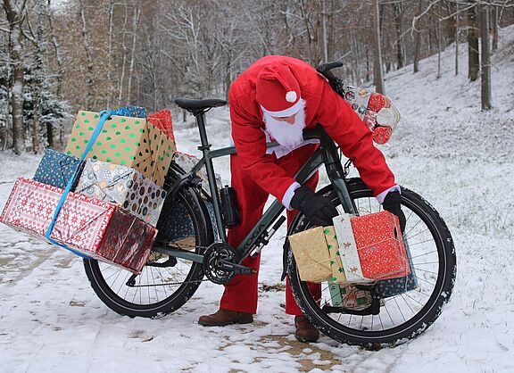 Man in Weihnachtskleidung beugt sich über ein mit Geschenken beladenes Fahrrad im Schnee. 